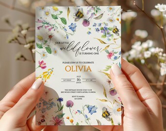 May include: A floral-themed first birthday party invitation for a girl named Olivia. The invitation features a watercolor design with wildflowers, bees, and butterflies on a white background. The text reads "Our little wildflower is turning one. Please join us to celebrate Olivia. Saturday, July 16, 20XX at 8 AM. The Heitman House Golf Club, 885 Wood Street, Fort Myers, Florida. RSVP to Maria, 123 123456."