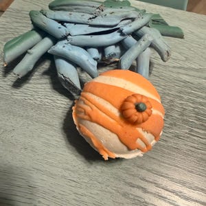 May include: A spherical bath bomb with a white base, orange stripes, and a small orange pumpkin decoration on top, positioned in front of a cluster of light blue painted wooden sticks arranged in a fan-like shape on a textured surface.