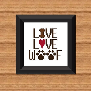 May include: A framed cross-stitch design with the words "LIVE LOVE WOOF" in brown letters. A brown bone is above the word "LIVE", a red heart is above the word "LOVE", and four brown paw prints are above the word "WOOF".