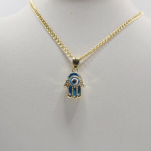 May include: A gold chain necklace with a hamsa hand pendant featuring a blue evil eye. The pendant is detailed with small gold beads around the edge.