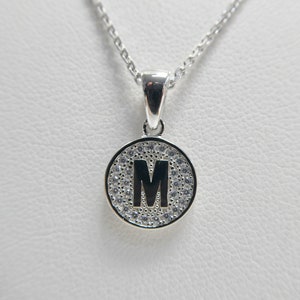 Puede incluir: Un collar de plata con un colgante redondo que presenta la letra "M" rodeada de pequeñas piedras transparentes.