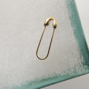 May include: A gold-toned safety pin with a smooth, polished finish. The pin features a classic U-shaped wire design with a rounded clasp mechanism at the top, showcasing a simple, functional form without any additional embellishments or decorative elements.