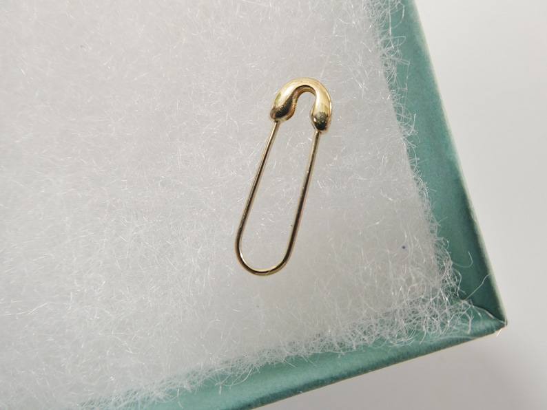 May include: A gold-toned safety pin with a smooth, polished finish. The pin features a classic U-shaped wire design with a rounded clasp mechanism at the top, showcasing a simple, functional form without any additional embellishments or decorative elements.
