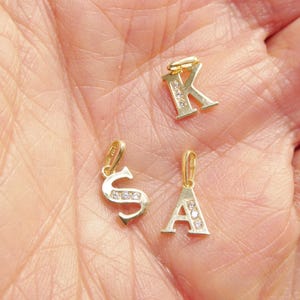 May include: Three gold-toned metal letter pendants featuring the letters K, S, and A. Each pendant has a small bail at the top and is embellished with a vertical row of three clear, sparkling stones set into the surface of the letter characters.