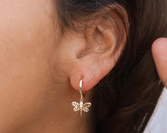 May include: Gold hoop earrings with a dangling dragonfly charm. The dragonfly is detailed with a delicate, openwork design.
