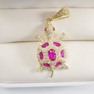 May include: A gold-tone turtle pendant featuring pink gemstones and clear cubic zirconia stones. The pendant is displayed on a white background.