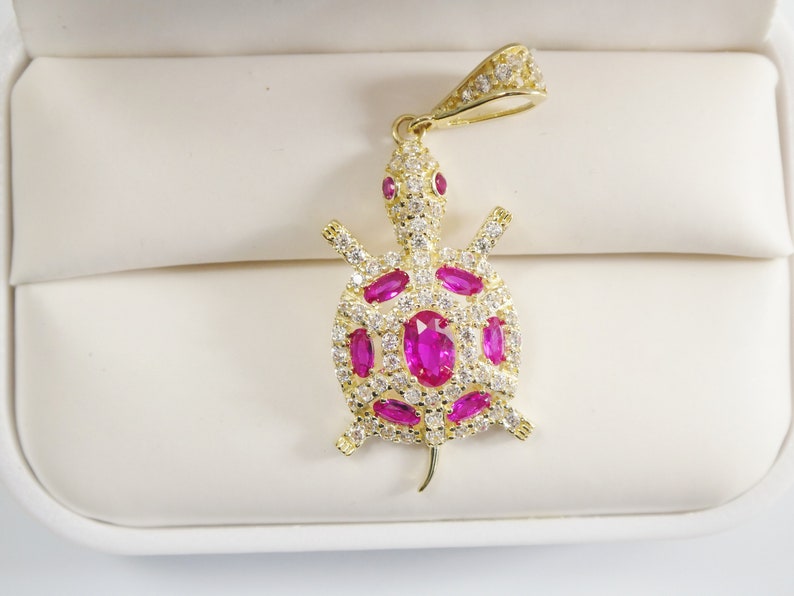 May include: A gold-tone turtle pendant featuring pink gemstones and clear cubic zirconia stones. The pendant is displayed on a white background.