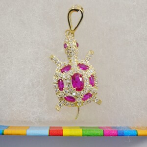 May include: A gold turtle pendant featuring pink and clear gemstones. The pendant has a loop at the top for attaching to a chain.