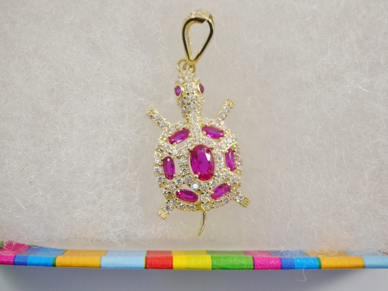 May include: A gold turtle pendant featuring pink and clear gemstones. The pendant has a loop at the top for attaching to a chain.