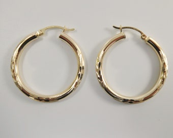 May include: A pair of gold hoop earrings. Each earring has a textured, patterned design around the outer surface. The earrings are round and have a clasp closure. The earrings are displayed on a white background.