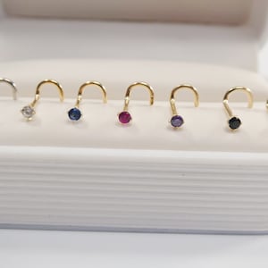 May include: A set of seven nose rings with different colored gemstones. The rings are made of gold and have a simple, curved design. The gemstones are clear, blue, pink, purple, and black.