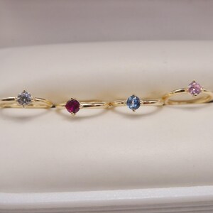 May include: Four gold rings with different colored gemstones: a light blue, a red, a blue, and a pink. The rings are displayed in a white jewelry box. The gemstones are round cut and set in a simple gold band.