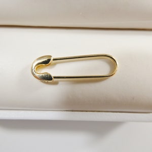 May include: A gold-toned safety pin with a smooth, polished finish. The pin features a classic elongated oval shape with a rounded clasp mechanism at one end and a thin, continuous wire frame that forms the body of the functional fastener.