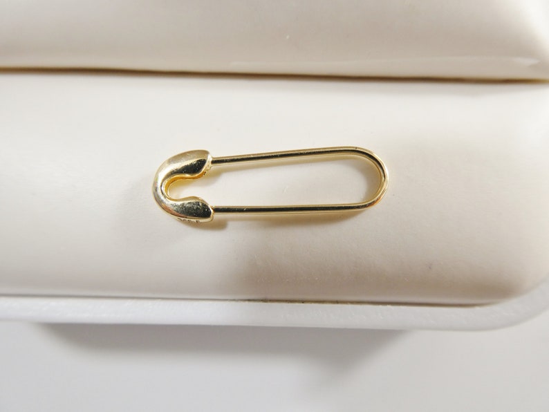 May include: A gold-toned safety pin with a smooth, polished finish. The pin features a classic elongated oval shape with a rounded clasp mechanism at one end and a thin, continuous wire frame that forms the body of the functional fastener.