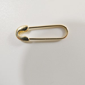 May include: A single gold-toned safety pin with a smooth, polished metallic finish. The pin features a classic U-shaped design with a rounded clasp mechanism at one end and a long, slender wire loop that extends to form the main body of the fastener.