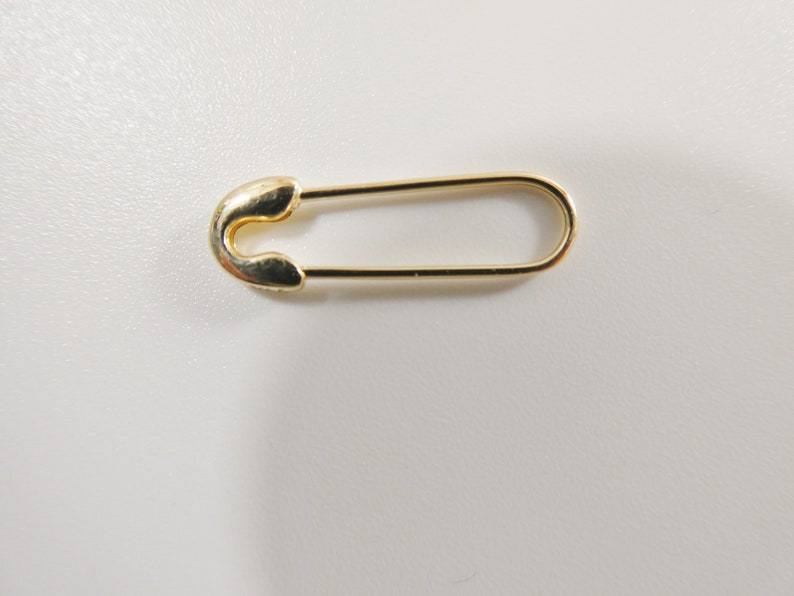 May include: A single gold-toned safety pin with a smooth, polished metallic finish. The pin features a classic U-shaped design with a rounded clasp mechanism at one end and a long, slender wire loop that extends to form the main body of the fastener.