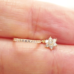 May include: A delicate gold earring featuring a small, sparkling flower pendant. The earring has a straight bar with small, clear stones and a flower-shaped charm with multiple clear stones. The earring is held in a person's hand.