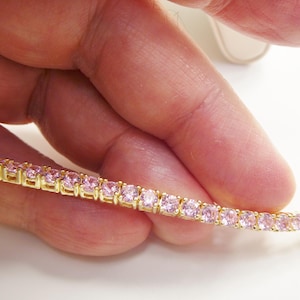 May include: A gold-toned bracelet with a row of pink gemstones. The gemstones are square-cut and set closely together. The bracelet is held by a hand against a white background.