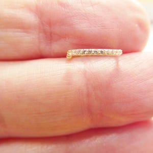 May include: A small, gold-colored ring with a row of small, clear gemstones. The ring is held between two fingers, showcasing its delicate design and sparkling details. The ring appears to be a piece of jewelry.
