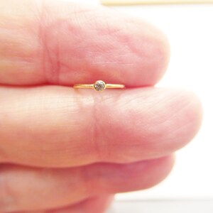 May include: A delicate gold ring with a small, round, clear gemstone set in the center. The ring is held between fingers, showcasing its minimalist design. The band appears thin and polished, highlighting the gemstone's sparkle.