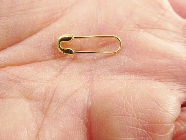 May include: A small, polished gold-tone safety pin with a smooth, metallic finish. The pin features a classic U-shaped clasp mechanism at one end and a long, slender wire body that extends to a rounded point, showcasing a simple, functional design.