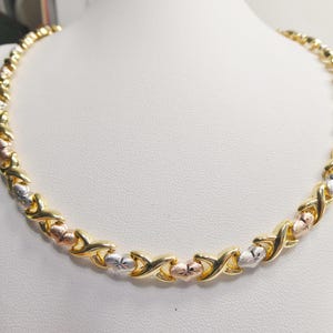 May include: A gold-tone necklace with alternating X and O links. The necklace features gold, rose gold, and silver-tone accents. The jewelry is displayed on a white bust.