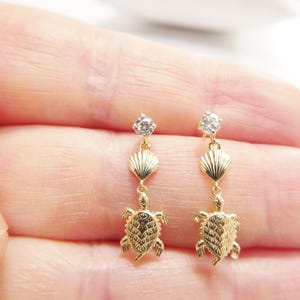 Puede incluir: Un par de pendientes de oro con un cristal transparente, una concha y un dije de tortuga.