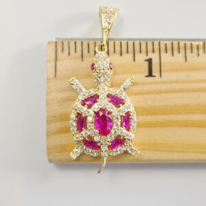 May include: Gold-tone turtle pendant with pink and white cubic zirconia stones. The pendant is approximately 2.5 cm long and is shown next to a ruler.