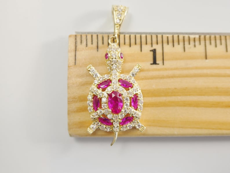 May include: Gold-tone turtle pendant with pink and white cubic zirconia stones. The pendant is approximately 2.5 cm long and is shown next to a ruler.