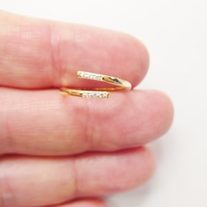 May include: A gold-coloured, open-ended ring with a wrap-around design. The ring features a row of small, clear gemstones on each end. The ring is shown against a neutral background.