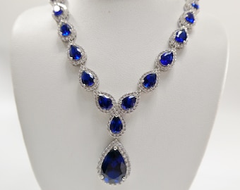 May include: A silver necklace with a teardrop-shaped pendant. The pendant is set with a large blue sapphire surrounded by smaller clear stones. The necklace is also set with smaller blue sapphires and clear stones.