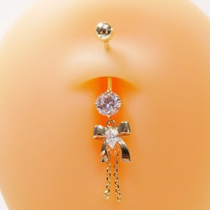 May include: Gold-toned belly button ring featuring a large, clear, round gem at the top and a dangling bow with a smaller, oval gem and chain accents. The jewelry is displayed on a light orange background.