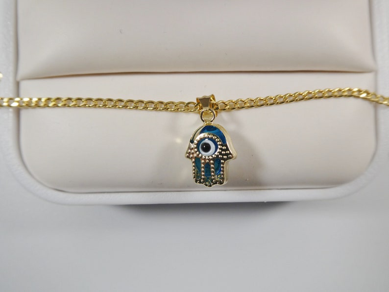 May include: A gold chain necklace with a hamsa hand charm. The charm is blue with a white eye in the center.