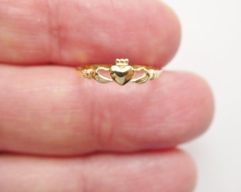 May include: A gold-colored Claddagh ring with a heart design and two hands on either side of the heart. The ring is worn on a finger.