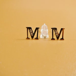 May include: A pair of gold-tone earrings with the letter "M" adorned with small clear stones. The earrings are on a light brown surface.