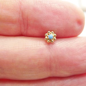 May include: A small, gold-colored flower-shaped stud earring with a light blue center. The earring is held between fingers, showcasing its delicate design. The earring's dimensions are not visible.