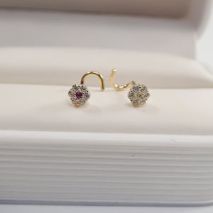 Puede incluir: Dos anillos de nariz de oro con una pequeña piedra roja en el centro de un diseño de flor rodeado de piedras transparentes.