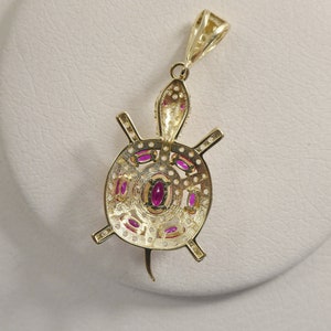 May include: Gold pendant necklace featuring a detailed turtle design with pink gemstones. The turtle's shell is intricately crafted with a circular pattern and the pendant hangs from a delicate chain.