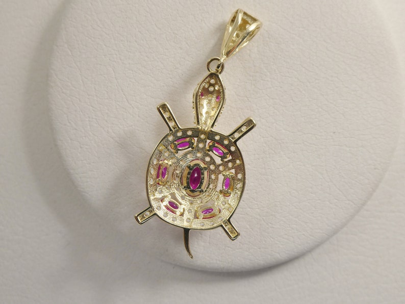 May include: Gold pendant necklace featuring a detailed turtle design with pink gemstones. The turtle's shell is intricately crafted with a circular pattern and the pendant hangs from a delicate chain.