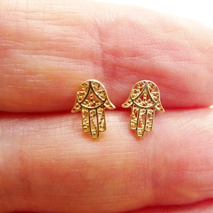 May include: A pair of gold-colored Hamsa hand stud earrings. Each earring features an openwork design with intricate details. The earrings are small and delicate, suitable for everyday wear.