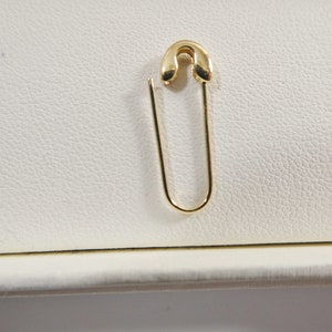 May include: A polished gold-tone safety pin earring featuring a curved top clasp and a smooth, U-shaped wire body. The metal has a reflective, high-shine finish, and the design mimics the classic shape of a functional safety pin in a jewelry format.