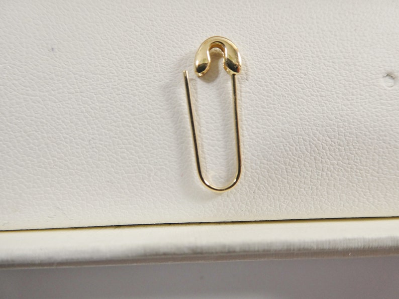 May include: A polished gold-tone safety pin earring featuring a curved top clasp and a smooth, U-shaped wire body. The metal has a reflective, high-shine finish, and the design mimics the classic shape of a functional safety pin in a jewelry format.