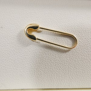 May include: A gold-toned safety pin with a smooth, polished metallic finish. The pin features a classic elongated oval shape with a rounded clasp mechanism at one end, designed to securely fasten fabric or other materials together in a functional manner.