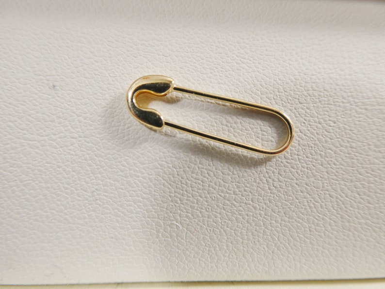 May include: A gold-toned safety pin with a smooth, polished metallic finish. The pin features a classic elongated oval shape with a rounded clasp mechanism at one end, designed to securely fasten fabric or other materials together in a functional manner.