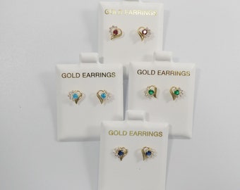 May include: Four pairs of gold heart-shaped earrings with different colored gemstones. Each pair is displayed on a white card with the text "GOLD EARRINGS". The gemstones are red, blue, green, and dark blue.