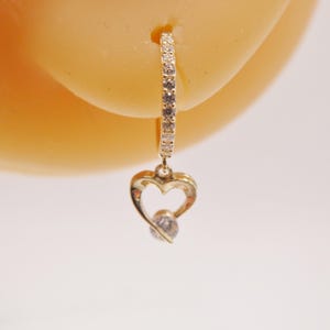 May include: A gold-colored earring with a heart-shaped charm. The earring features a small hoop lined with clear stones and a heart pendant with a single, clear stone at its center. The earring is shown against a light background.