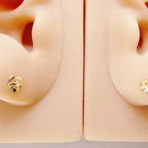 May include: A pair of small, gold-colored heart-shaped stud earrings. The earrings have a textured surface and are displayed on a light-colored mannequin head. The earrings are a classic jewelry item.