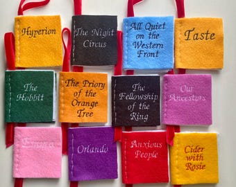 May include: A collection of miniature felt book ornaments in various colors, each with a title embroidered in white. Titles include "Hyperion," "The Hobbit," and "Orlando." Red ribbon loops are attached for hanging.