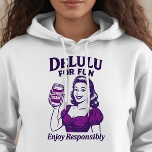 Puede incluir: Sudadera con capucha blanca con un gráfico que presenta una ilustración de estilo vintage de una mujer sosteniendo un tarro. El texto en la sudadera con capucha dice "DELULU FOR FUN" y "Enjoy Responsibly" en morado. La sudadera tiene una capucha con cordón y un bolsillo delantero.