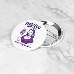 May include: White button featuring a retro illustration of a woman holding a drink, with the text "DELULU FOR FUN" and "Enjoy Responsibly" in purple. The button has a silver pin back.